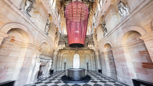 A Hall with a black and white checked floor and stone walls. Suspended from the ceiling is a large installation with long red fabric strips that connect at a central point.